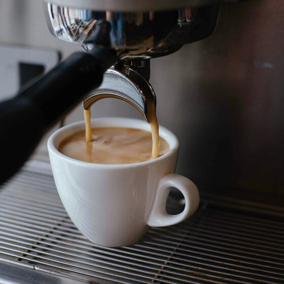 Nahaufnahme einer Kaffeemaschine, die frischen Espresso in eine weiße Keramiktasse gießt, die auf der Metallabtropfschale der Maschine steht.