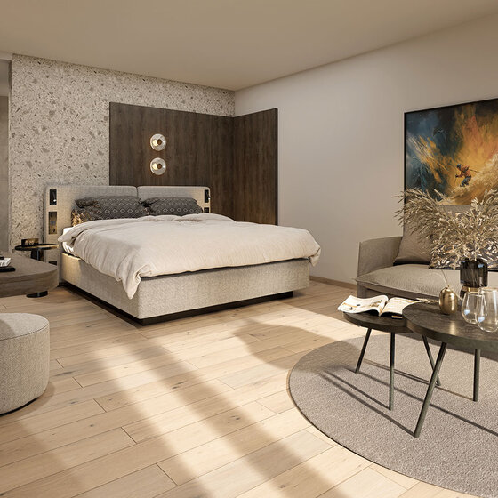 Ein modernes Schlafzimmer mit einem hellen Holzboden, einem Doppelbett, einem grauen Sofa, runden Couchtischen, einem Hocker, Wandkunst und dekorativen Pflanzen. Neutrale Töne und minimalistisches Dekor schaffen eine gemütliche, stilvolle Atmosphäre.