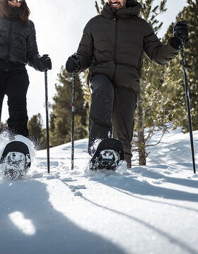 Zwei Personen in Schneeschuhen und Winterkleidung gehen durch frischen Schnee in einem sonnenbeschienenen Wald, wobei sie Stöcke zur Unterstützung benutzen. Im Hintergrund sind hohe Kiefern und ein klarer Himmel zu sehen.