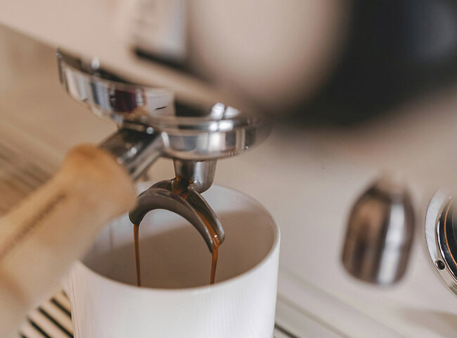 Nahaufnahme einer Espressomaschine, die Kaffee zubereitet, wobei dunkler Kaffee aus dem Auslauf des Siebträgers in eine weiße Keramiktasse fließt.