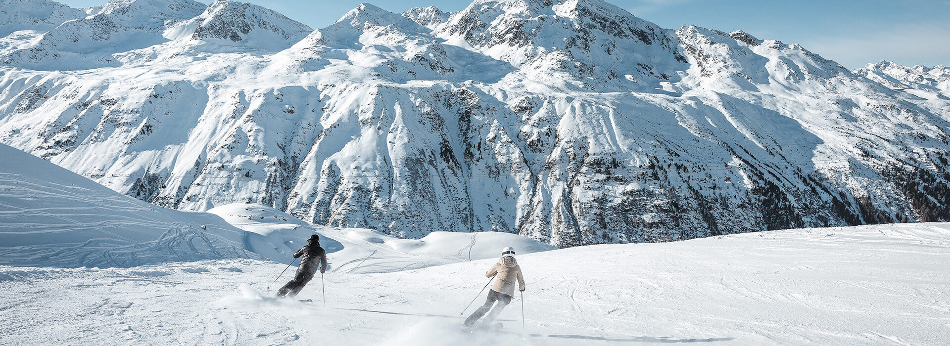 Zwei Personen fahren auf Skiern einen verschneiten Hang hinunter, mit hohen, schroffen, schneebedeckten Bergen und einem blauen Himmel im Hintergrund.