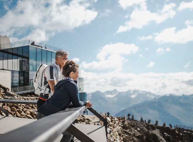 Zwei Personen stehen an einem Geländer mit Blick auf die Berge und ein modernes Gebäude unter einem teilweise bewölkten Himmel.