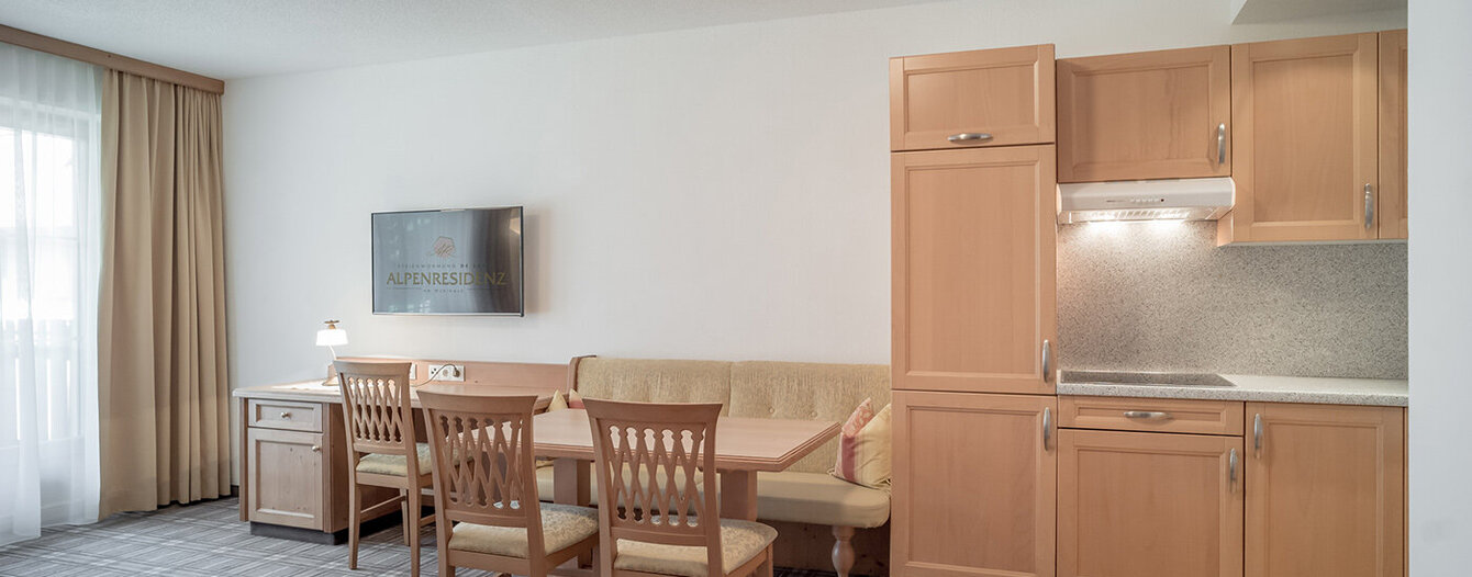 Eine helle, moderne Küche und ein Essbereich mit hellen Holzschränken, einem Esstisch mit vier Stühlen, einer eingebauten Bank, einem Schreibtisch und einem an der Wand montierten Fernseher. Ein großes Fenster mit Vorhängen lässt viel natürliches Licht herein.