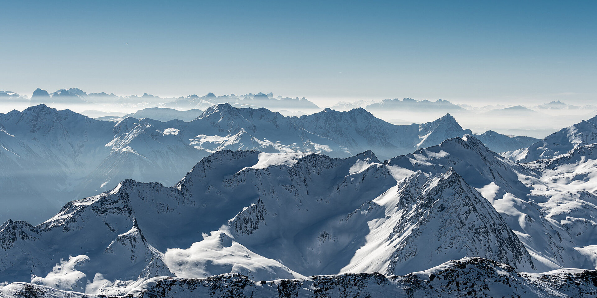 Schneebedeckte Berggipfel erstrecken sich in der Ferne unter einem klaren blauen Himmel und sanftem Morgenlicht.