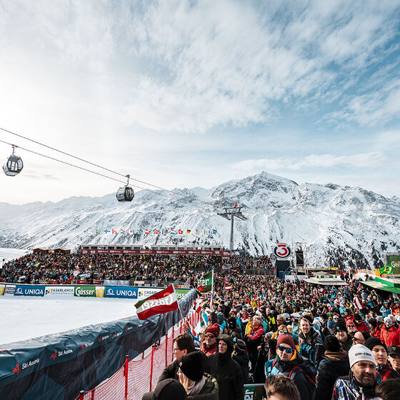 Eine große Menschenmenge versammelt sich in einem verschneiten Skigebiet, mit Bergen und Skiliften im Hintergrund. Die Zuschauer stehen in der Nähe der Skipiste, viele tragen Winterkleidung bei teilweise bewölktem Himmel.