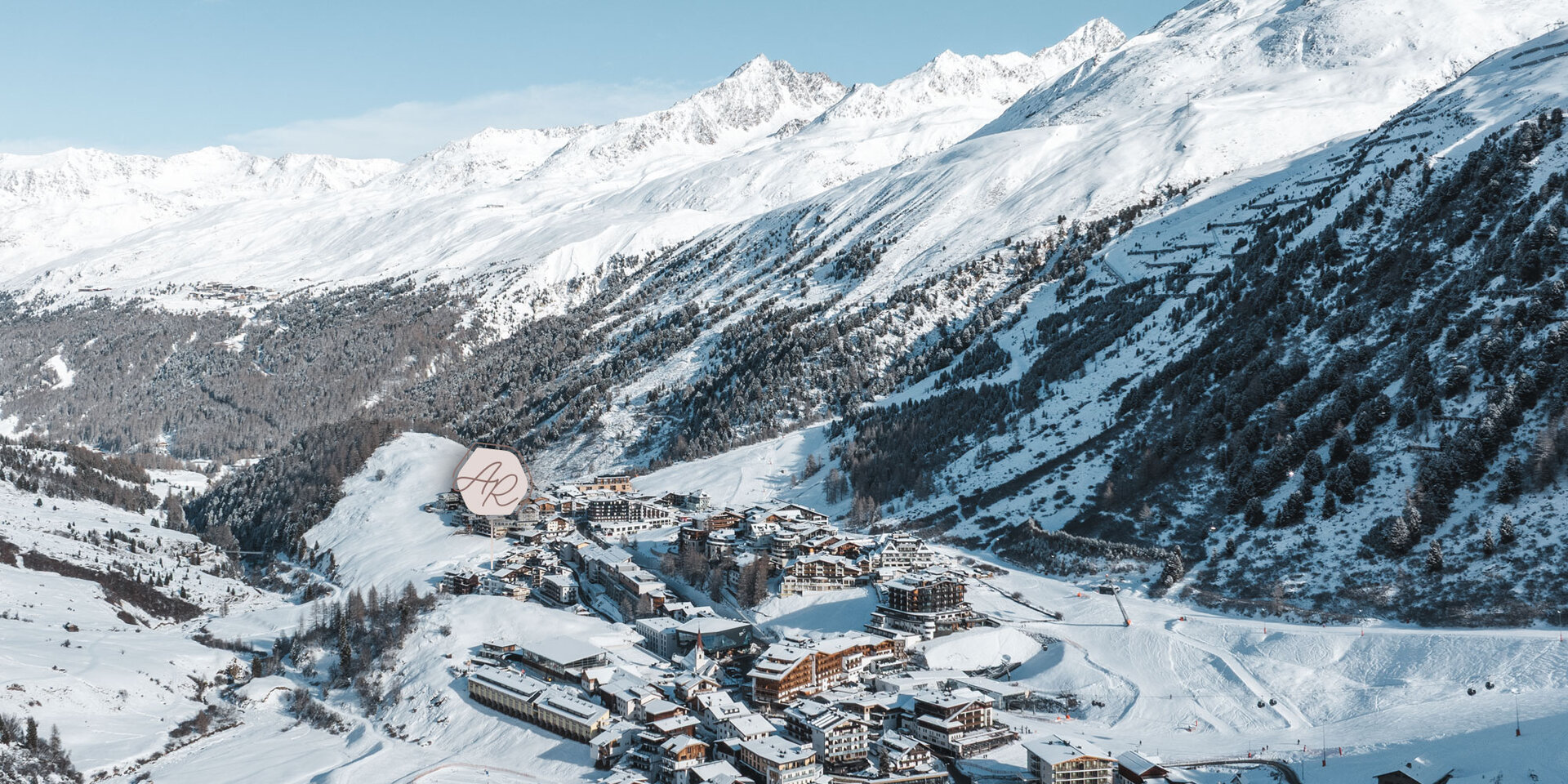 Luftaufnahme eines schneebedeckten Alpendorfs, umgeben von Bergen und Wäldern, mit klarem, blauem Himmel und fernen schneebedeckten Gipfeln im Hintergrund. Das Logo der Alpenresidenz wurde im Bild dort platziert, wo die Alpenresidenz in Gurgl steht.