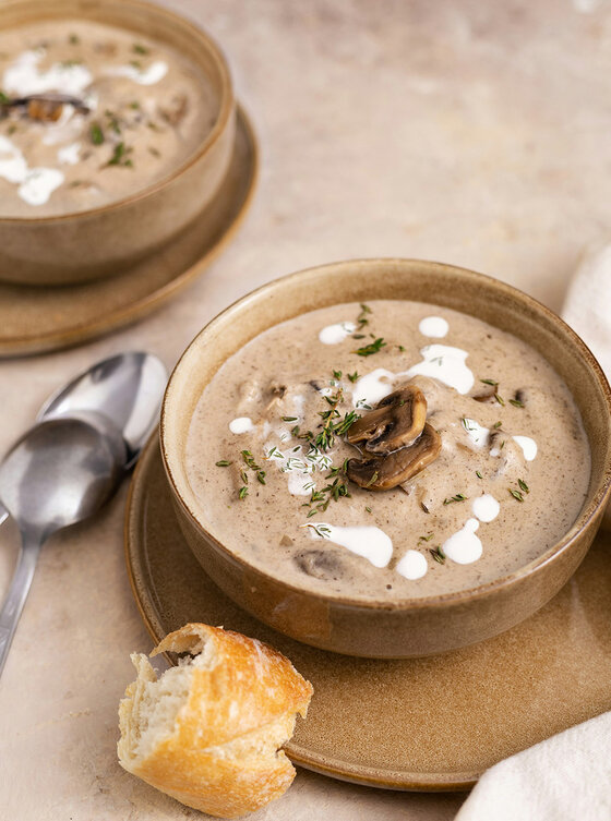 Eine Schüssel mit cremiger Pilzsuppe, garniert mit Pilzscheiben, Sahne und Kräutern, serviert auf einem braunen Teller mit einem Stück Brot und einem Löffel daneben. Im Hintergrund steht eine weitere Schüssel mit Suppe.