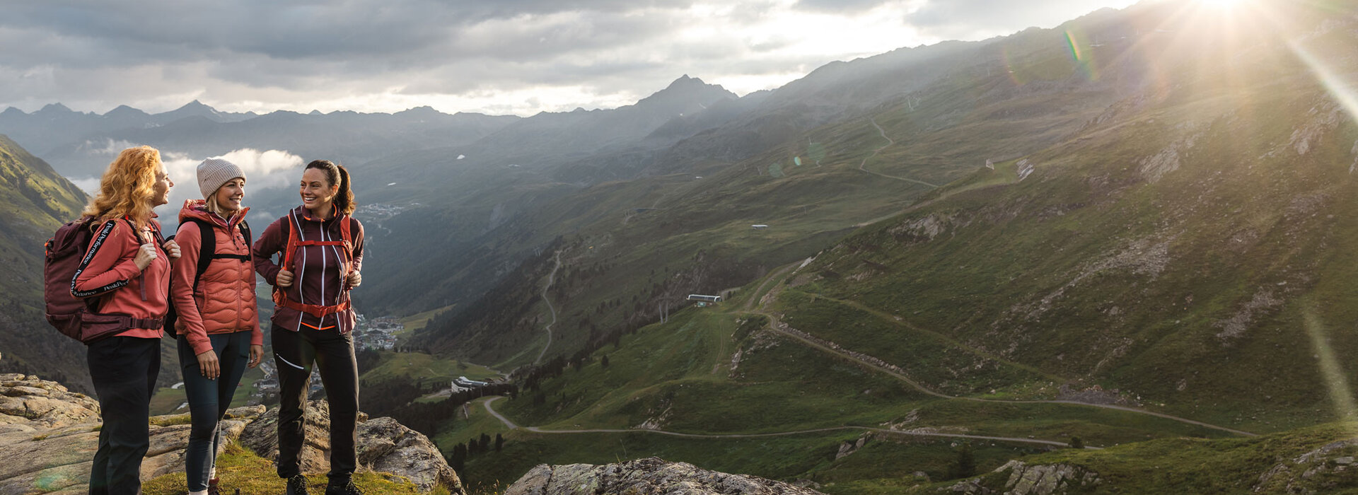Drei Frauen mit Rucksäcken und Outdoor-Jacken stehen auf einer felsigen Bergkuppe, lächeln und unterhalten sich, mit einer malerischen Berglandschaft und der untergehenden Sonne im Hintergrund.