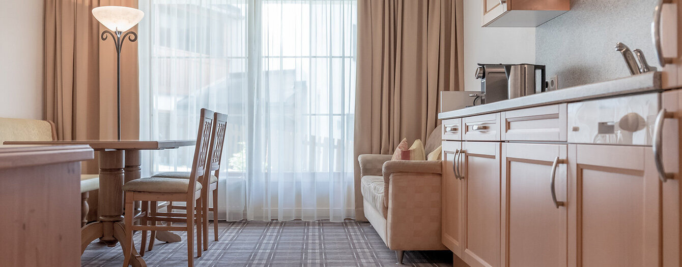 Eine helle, gemütliche Küche und ein Essbereich mit Holzschränken, einem Esstisch mit Stühlen, einem beigen Sofa, einer Stehlampe und großen Fenstern mit Vorhängen, die natürliches Licht hereinlassen.