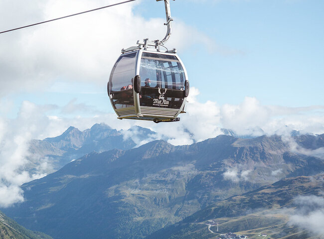 Eine Seilbahn fährt über grüne Berge und Täler unter einem blauen Himmel mit Wolken.